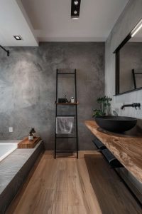 Modern minimalist bathroom with wooden flooring, a black ladder shelf, wall-mounted mirror, black basin, and concrete walls. A bathtub is partially visible on the left.