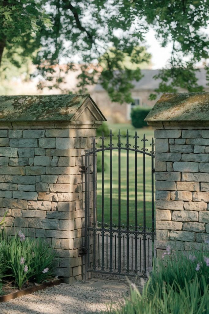 A wrought iron gate set within a stone wall, leading to a grassy garden area, surrounded by green plants and trees.