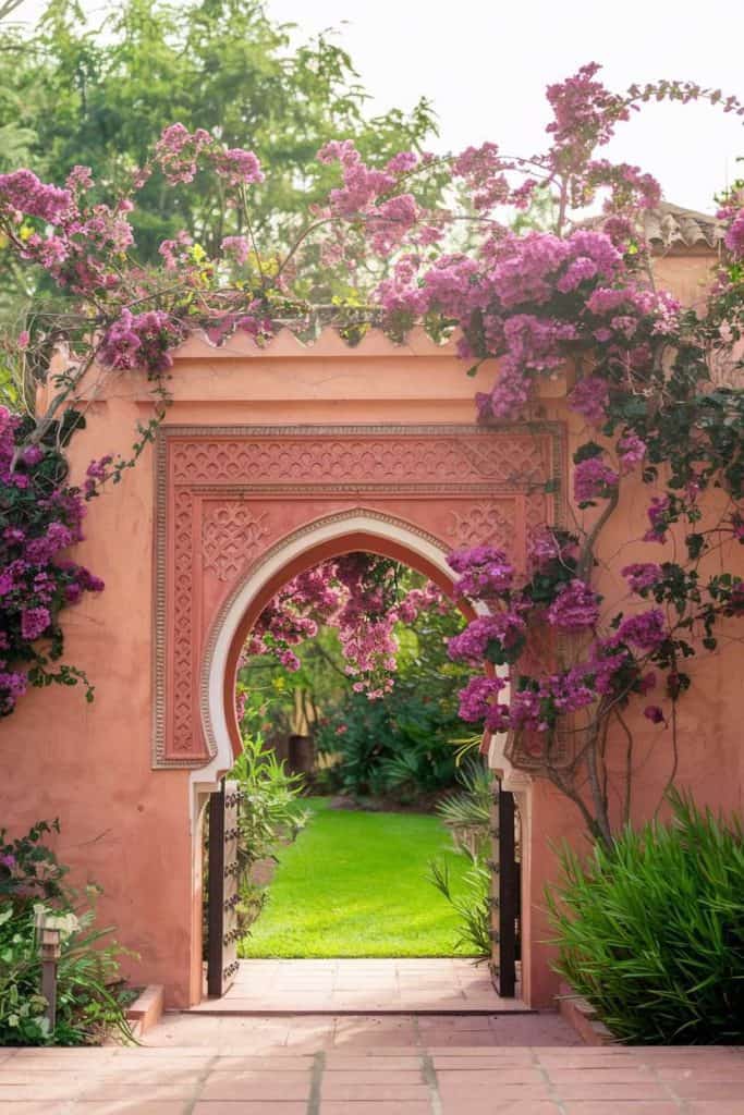 An ornate archway adorned with purple bougainvillea leads to a lush garden with a green lawn.