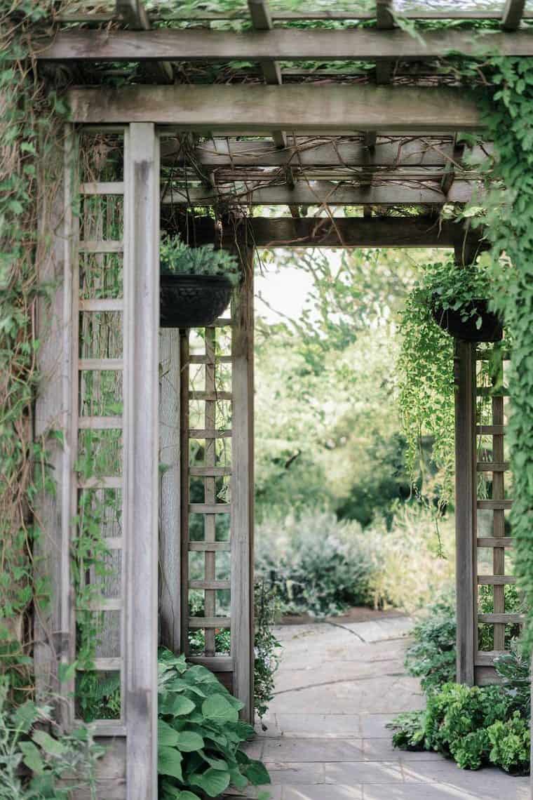 Wooden garden archway covered with green vines, leading to a stone path and lush greenery beyond.