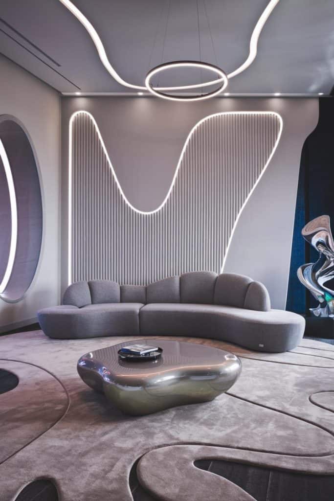 Modern living room with a gray, curved sofa, abstract wall lighting, a metallic coffee table, and textured rugs. The design features contemporary and futuristic elements.