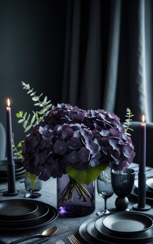 A dining table set with black plates, dark cutlery, two lit purple candles, and a centerpiece of purple hydrangeas in a vase, against a dark background.