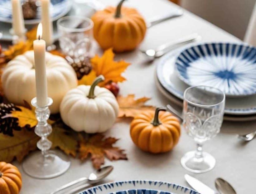 A dining table set with blue and white plates, silver cutlery, glasses, and decorated with small pumpkins, white candles, and autumn leaves.