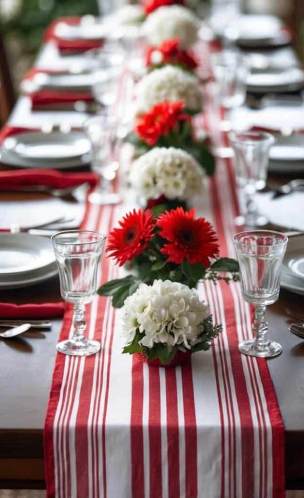 A long dining table set with white plates, glasses, and red napkins, decorated with red and white flowers on a red and white striped table runner.