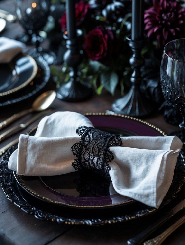 A formal dining table set with black plates, gold cutlery, a white napkin with a black lace ring, and black candles, with dark flowers in the background.