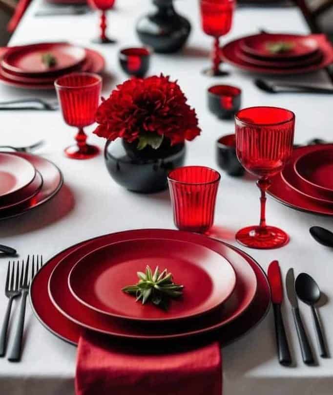 A long table set with red plates, glasses, and napkins on a white tablecloth, decorated with red flower centerpieces in black vases.