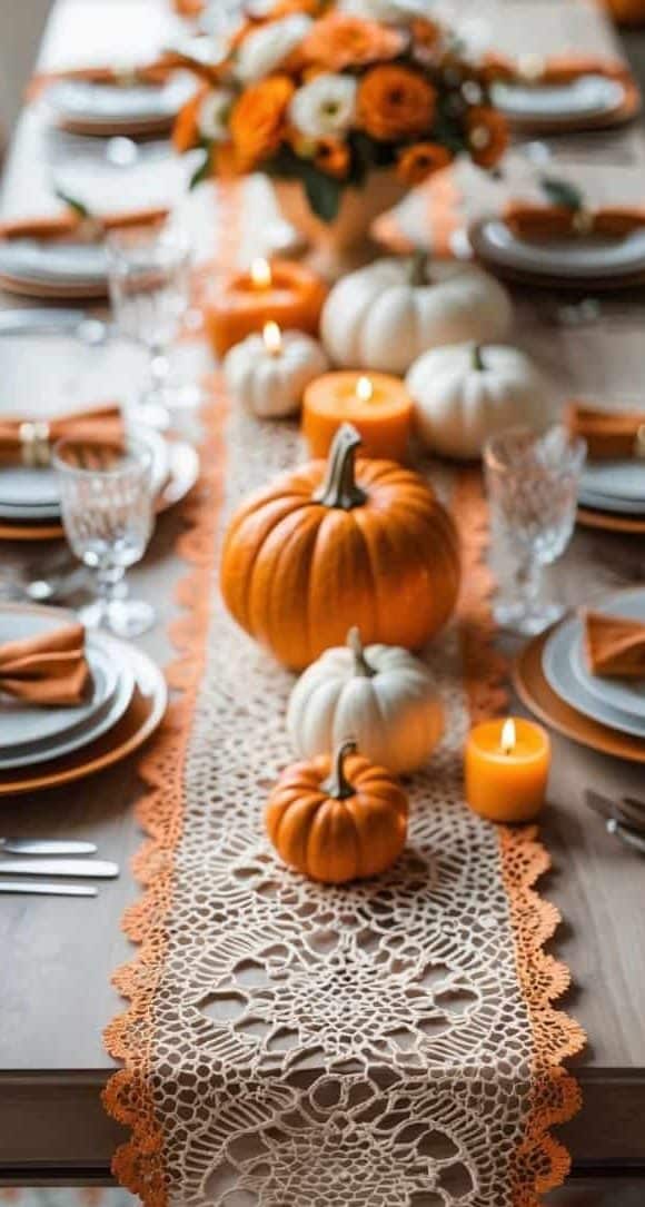 A dining table set with plates, glasses, orange napkins, a lace table runner, white and orange pumpkins, candles, and a floral centerpiece.