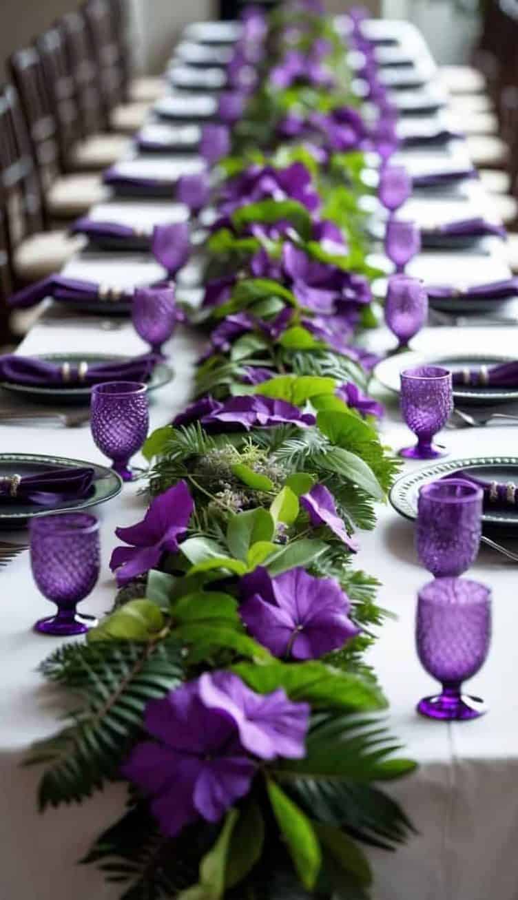 A long dining table is decorated with a green and purple floral garland, matching purple napkins, and purple glassware.
