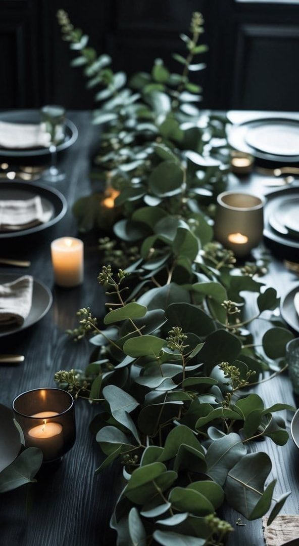A dark wooden dining table set with gray plates, beige napkins, gold cutlery, lit candles, and a green eucalyptus garland as a centerpiece.