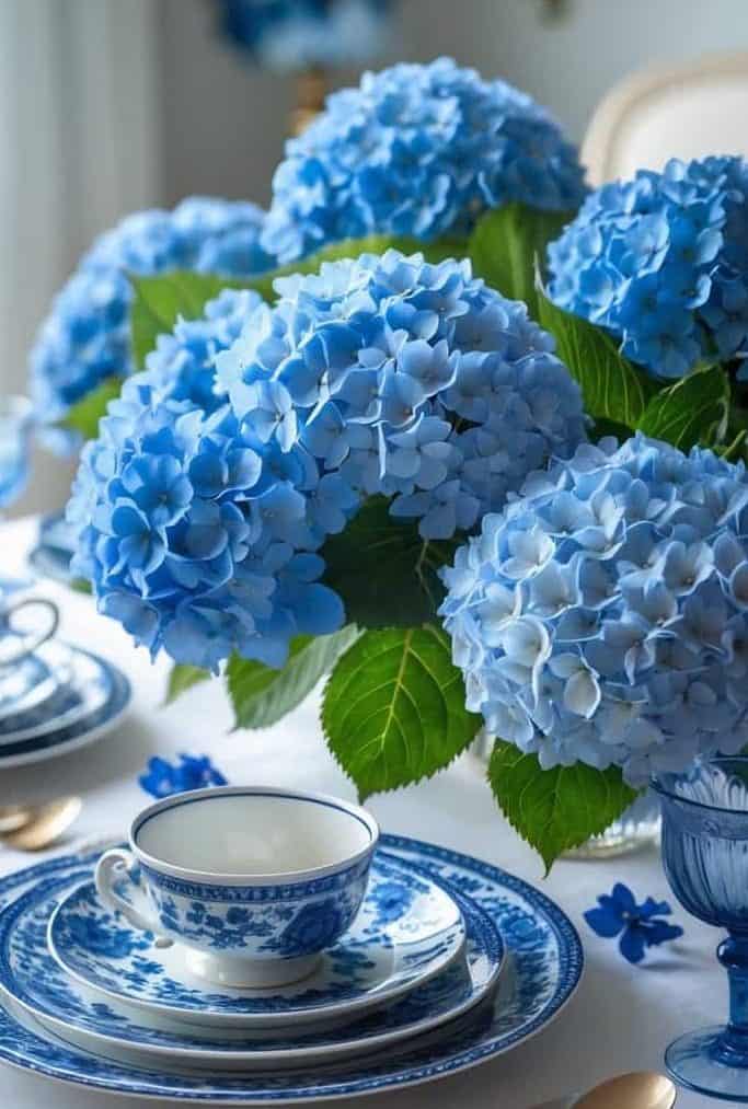 A dining table set with blue and white china and teacups, featuring a centerpiece of large blue hydrangea flowers.