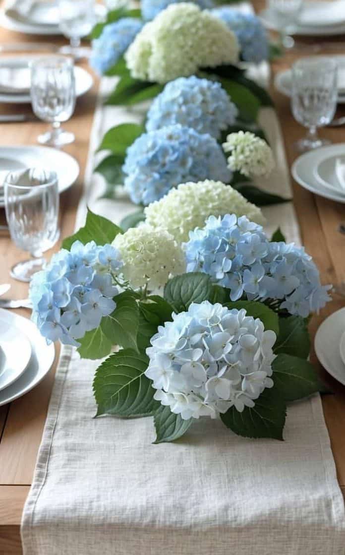 A wooden dining table set with white plates, silverware, and glasses, featuring a centerpiece of blue and white hydrangea flowers on a cream table runner.