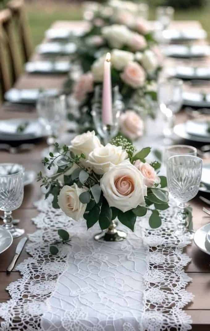 A wooden table set for a formal meal features white lace runners, glassware, plates, silverware, and floral centerpieces with white and pale pink roses.