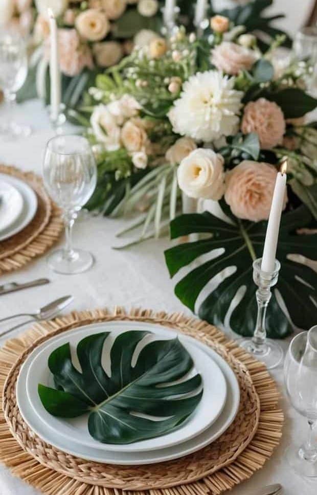A formal dining table is set with white plates on woven placemats, decorated with green leaves. There are floral centerpieces, wine glasses, and tall white candles.