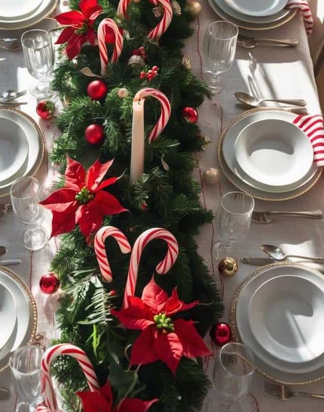 A dining table set for six with white dishes, gold utensils, and glasses, decorated with evergreen garland, red poinsettias, candy canes, and tall candles in a festive holiday theme.