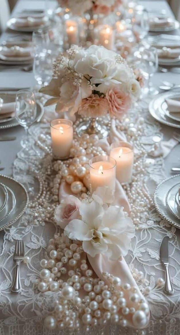 Elegant dining table set with white plates, silver cutlery, lit candles, pearl decorations, and white floral centerpieces on a lace tablecloth.