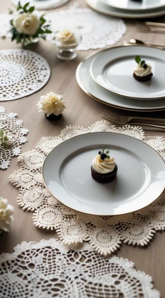 A neatly set dining table with white plates, gold-rimmed chargers, and a single cupcake on each plate, decorated with lace placemats and small white flowers.