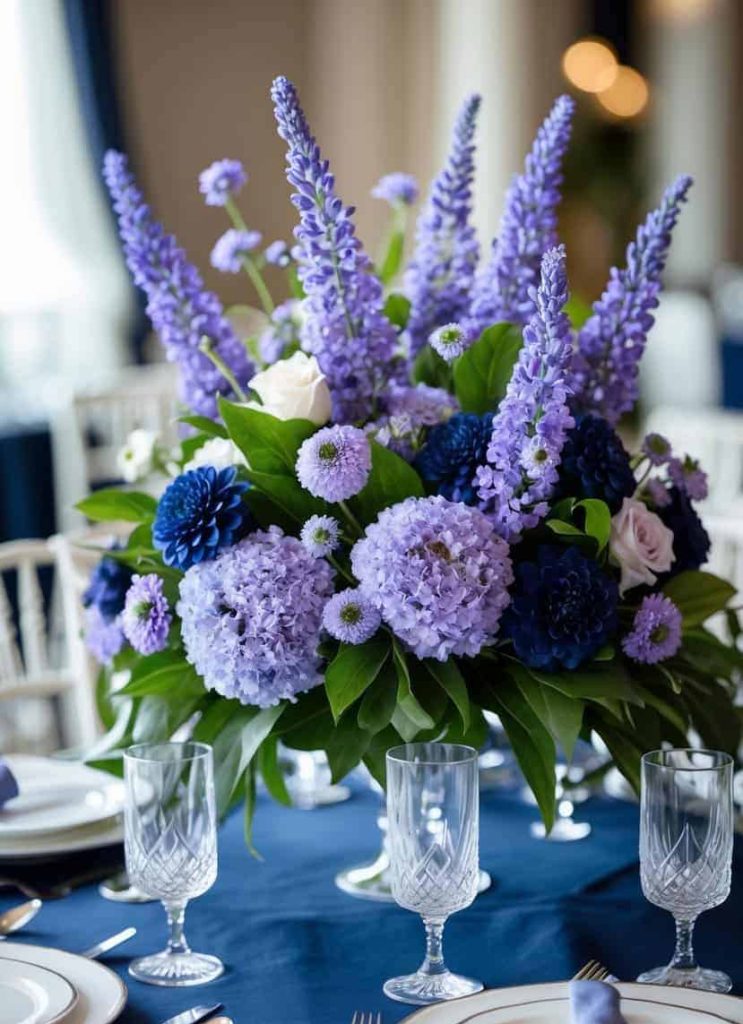 A centerpiece with purple delphiniums, hydrangeas, dark blue dahlias, and light roses sits on a table set with plates, glasses, and silverware.