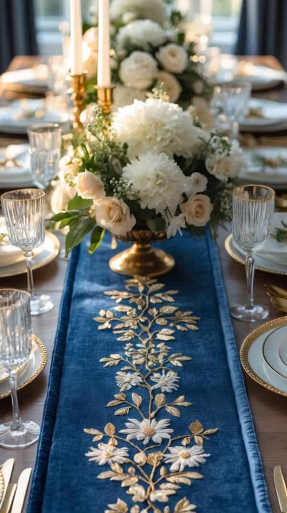 A formal dining table set with white and gold dishes, glassware, white floral centerpieces, and a blue table runner with gold floral embroidery.
