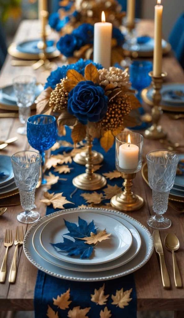 A dining table set with blue and gold-themed decorations, including candles, glassware, artificial flowers, and scattered ornamental leaves.