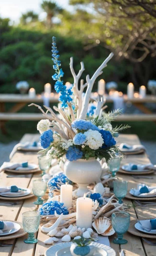A rustic picnic table is set outdoors with blue and white floral centerpieces, candles, seashells, and blue-tinted glassware, creating a coastal-themed dining arrangement.