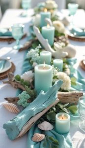 A table centerpiece with blue candles, driftwood, seashells, white flowers, and greenery arranged on a blue cloth, set for a meal with aqua glassware.