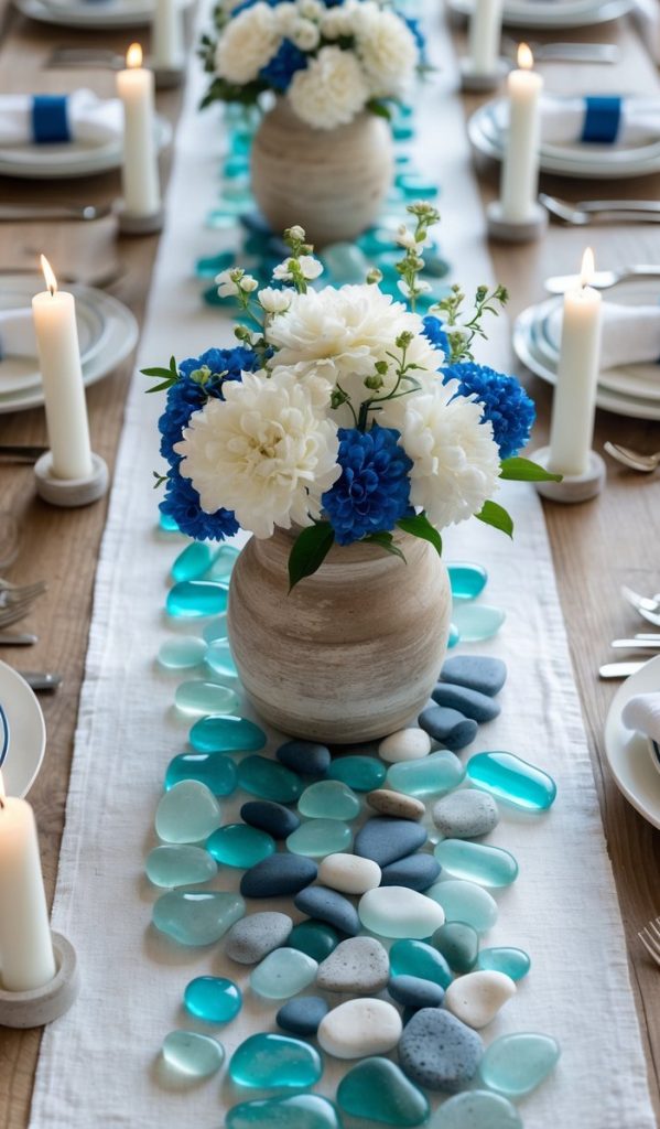 A dining table set with white plates, blue napkins, lit white candles, and vases of white and blue flowers on a runner decorated with blue and turquoise stones.